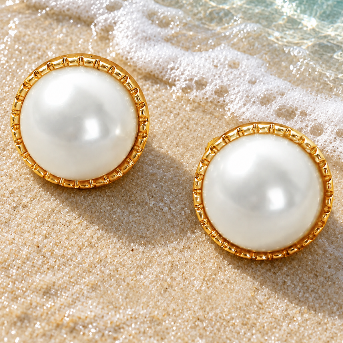A pair of round pearl stud earrings, resting on sandy beach ground 