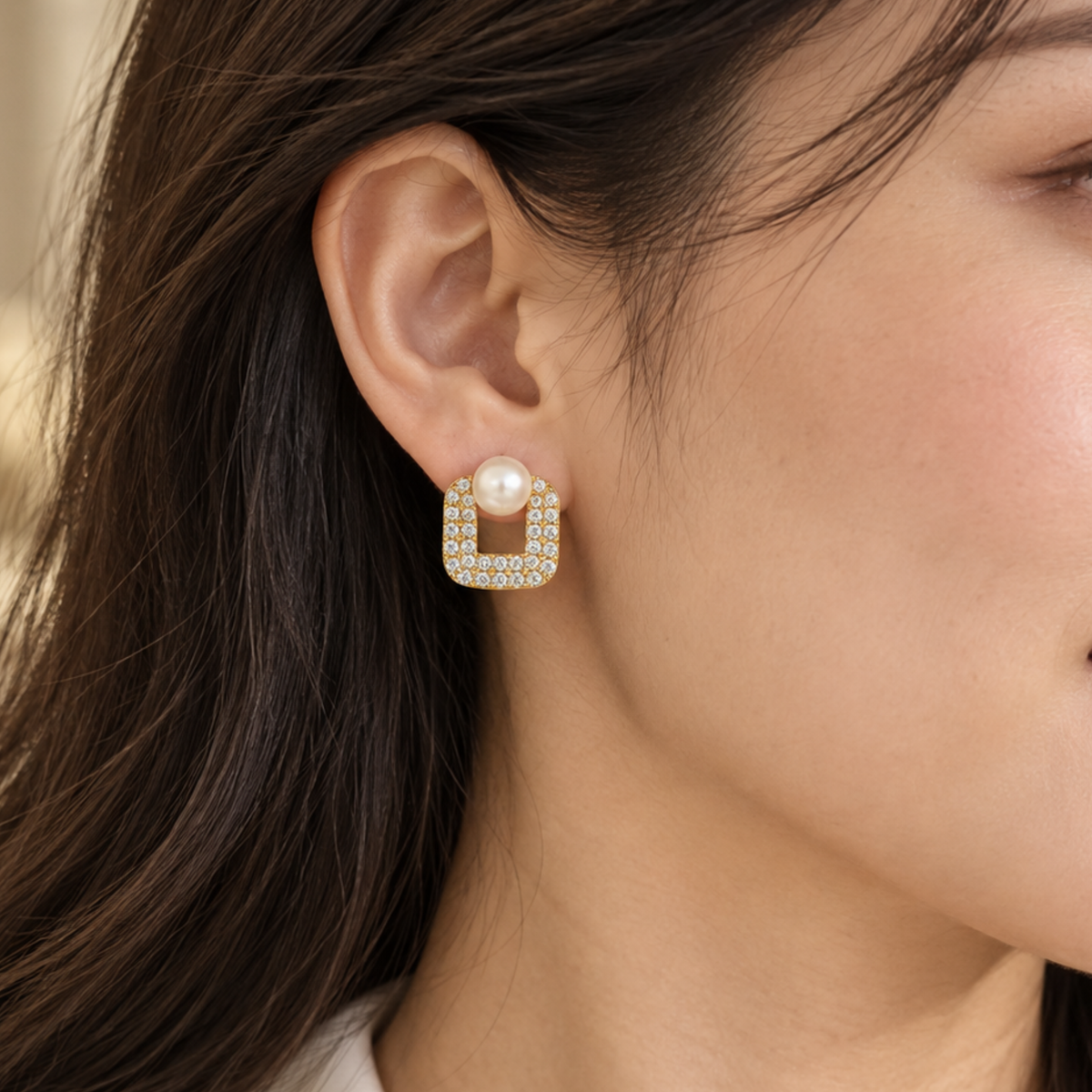 A woman wearing a gold square stud earring with a central white pearl 